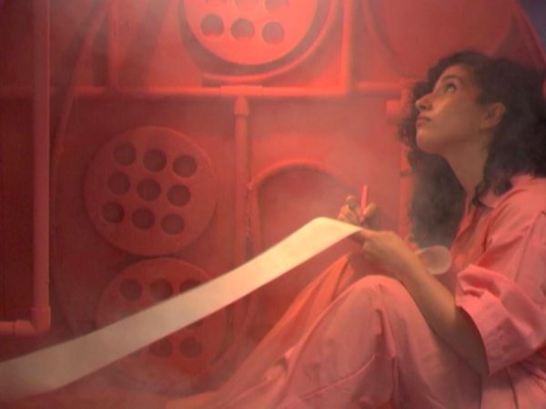 A film still shows a woman sitting on the floor, dressed in the same pink color as the wall behind her and writing on a long sheet of paper