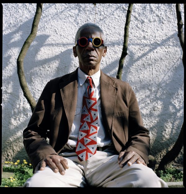 A photographic portrait shows a seated, dark-skinned man wearing a brown jacket, colorful sunglasses, and a boldly patterned tie.