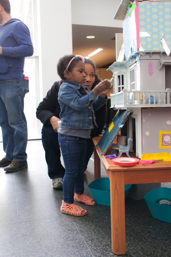 A woman crouches next to a young girl playing with a large doll house in the MCA's atrium.