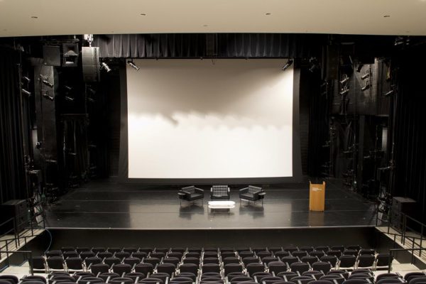 A large white screen hangs above three chairs and a table set up on a black stage in an empty theater venue