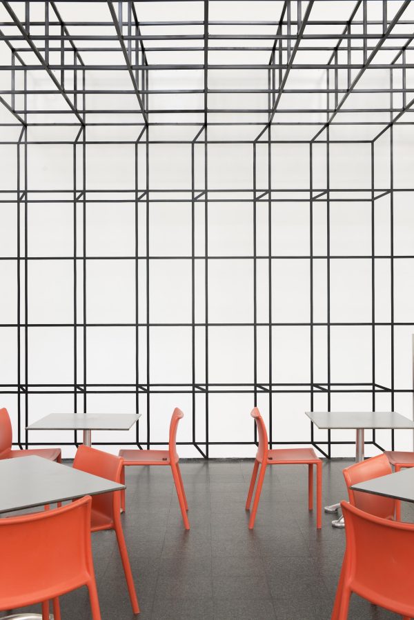A room with orange chairs and white tables and a ceiling plane made of gray, open-sided cubes that seamlessly transitions into a grid of illusionistic cubes on the wall