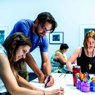 Three adults drawing on a table with framed artworks in the background.