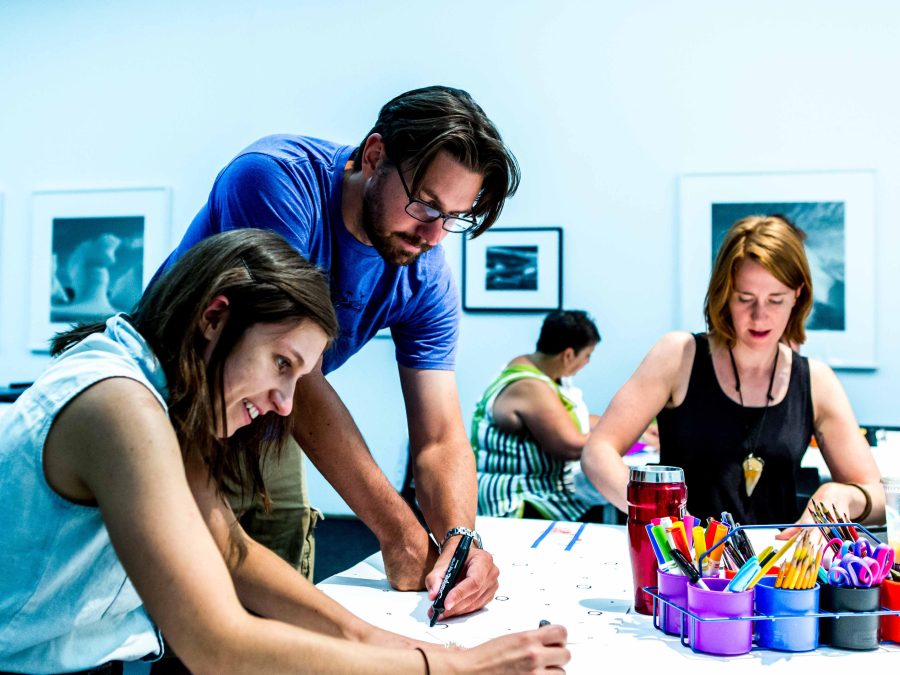 Three adults drawing on a table with framed artworks in the background.