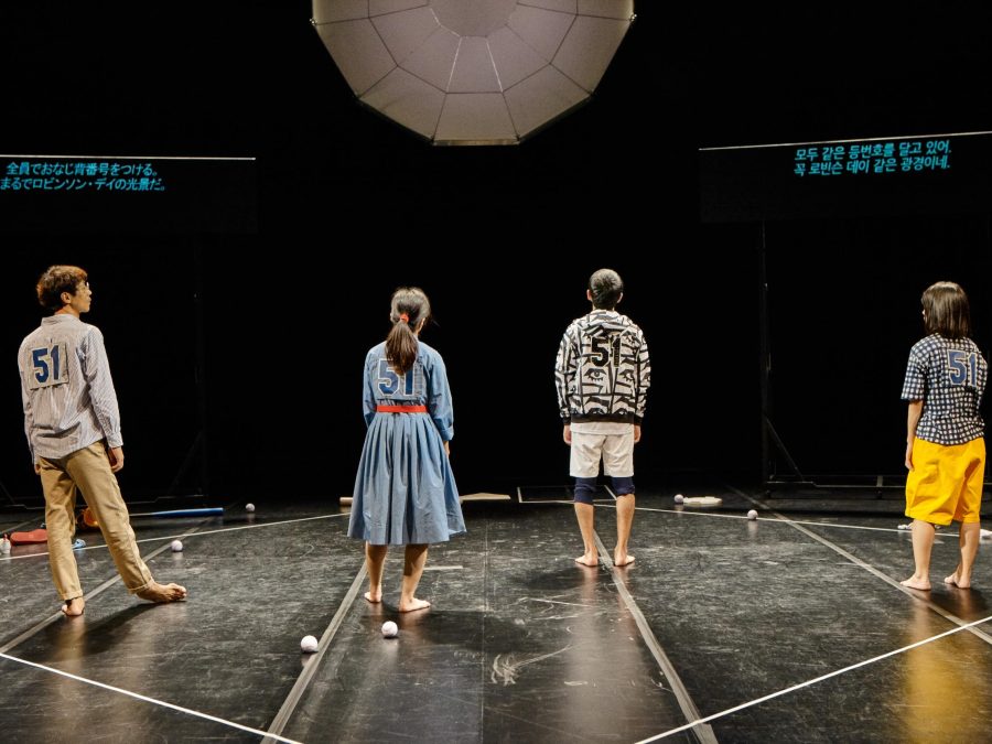 A performance still shows four people wearing the number 51, facing the back of the dark stage, and standing inside the white outline of a square with a few baseballs at their feet