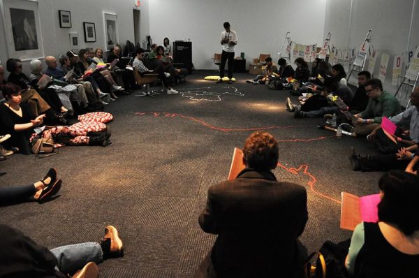 A group of people sit in a circle on a floor in a dimly lit room