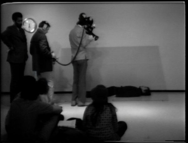 A camera crew films Chris Burden lying on the floor under a piece of glass