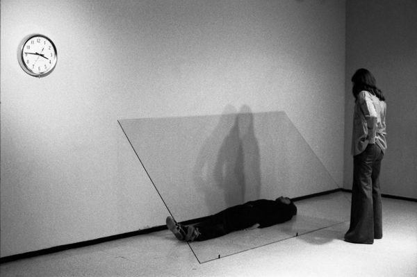 A person observes a man lying on the floor underneath a sheet of glass.