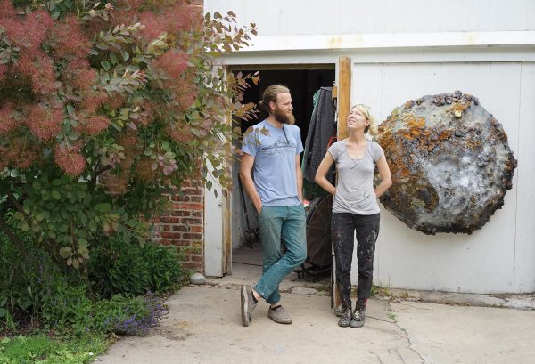 Two people with ivory skin stand outside a white building featuring a large half circle sculpture resembling a grey moon or planet