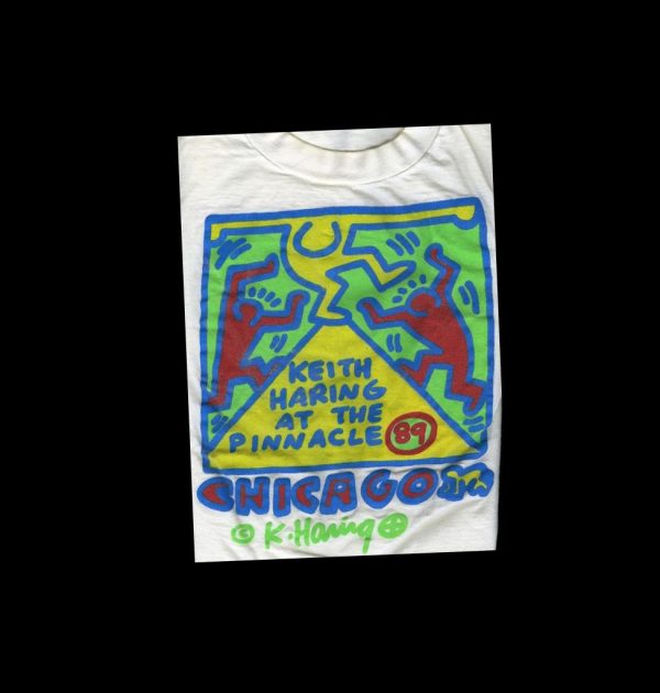 On a white tee shirt, three colorful cartoon figures dance on a yellow triangle inside a blue-outlined square with light green fill.