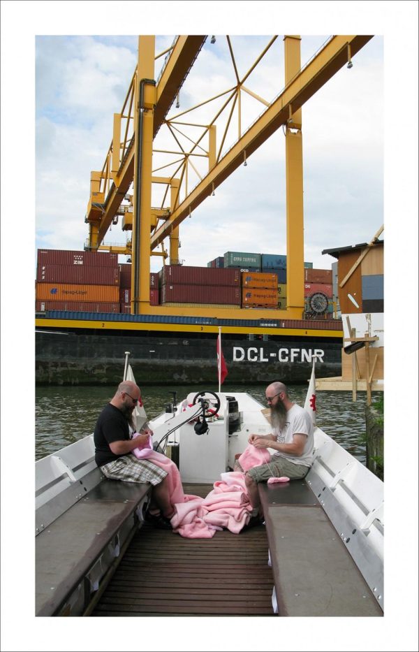 Two people knit a pink object on a boat near an industrial crane