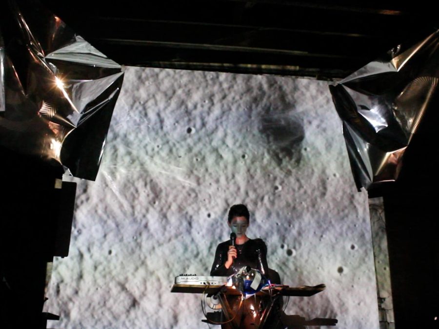 A person holding a microphone sits at an unusual desk with a large black-and-white backdrop that resembles the cratered surface of the moon. Two crinkled metallic sheets are suspended above.