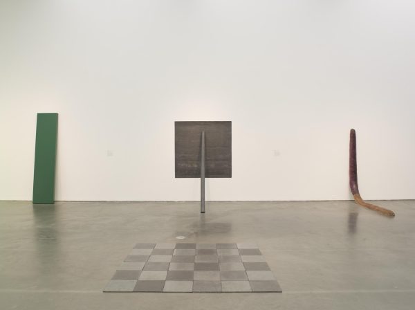 Installation view of four minimalist sculptures including a green rectangular plank and a worm-like form leaning against the wall, a square grey sheet held to the wall by a pipe, and on the ground, a square grid made of dark and light grey squares