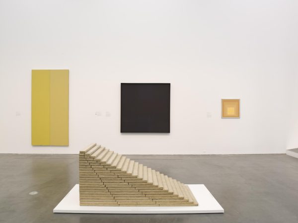A gallery view featuring artist Jackie Ferrara's Stairway, a staircase-like sculpture made out of wood in the foreground, and additional color-field paintings in background.