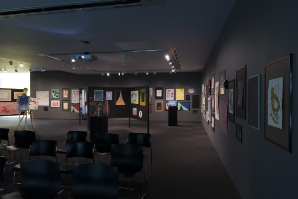 Installation view of paintings and sculptures in a grey gallery, rows of chairs in the foreground, and a humanoid figure on the left standing at an easel and canvas