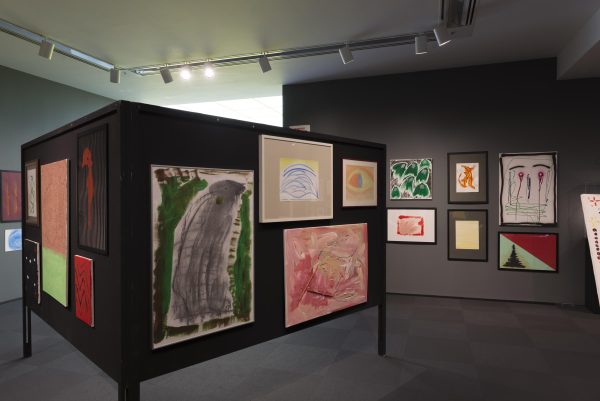 Colorful, abstract paintings hung in clusters on the grey walls of a gallery and on a free-standing folding board