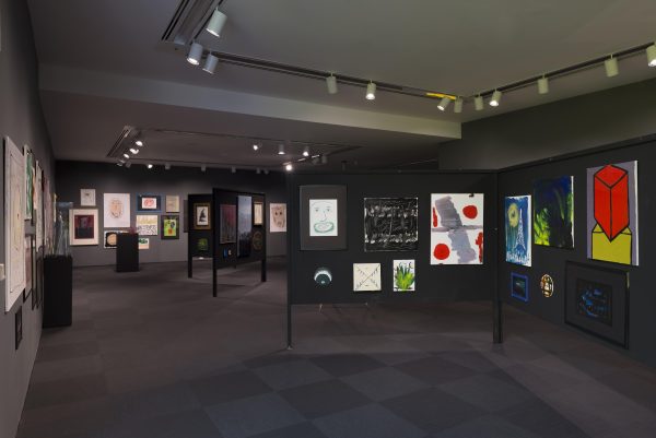 Colorful, abstract paintings hung in clusters on the grey walls of a gallery and an abstract sculpture on a pedestal