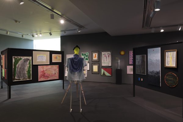 A humanlike figure with a round lime green head and two spindly legs wears a plaid shirt and stands in front of a tripod, facing an array of drawings and paintings hung on a zigzagging black display board.
