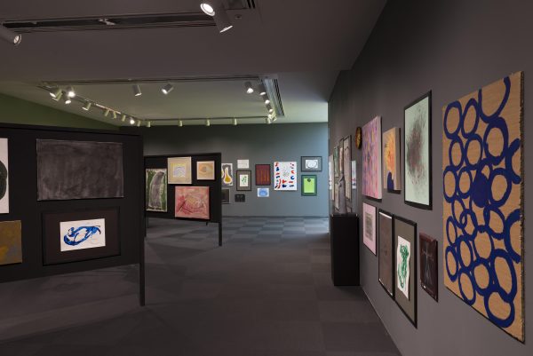 More than two dozen colorful abstract paintings are hung in clusters on gray walls and black folding display boards