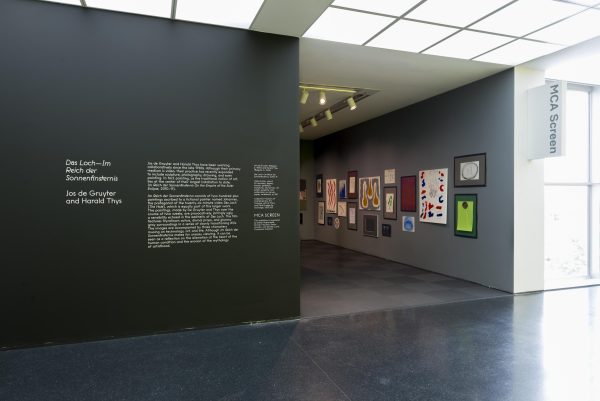 The entrance to an MCA Screen exhibition hosts white gallery text and colorful paintings hung in clusters on the opposite wall.