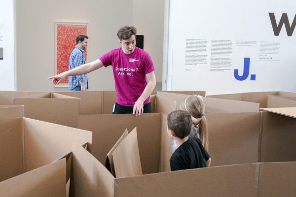 Two children in a cardboard maze look at man pointing to our left.