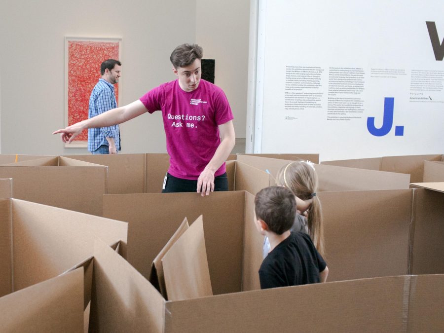 Two children in a cardboard maze look at man pointing to our left.