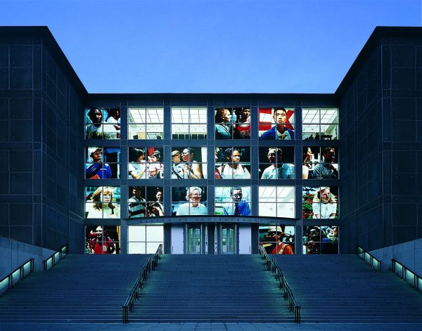 The front windows of the MCA illuminated with large close-up photographs of people.