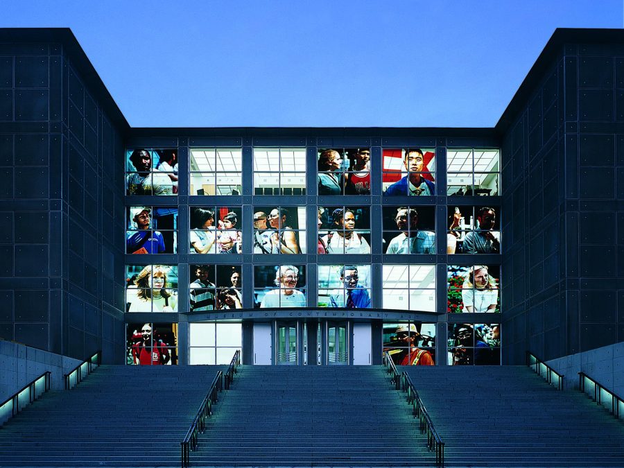 The front windows of the MCA illuminated with large close-up photographs of people.