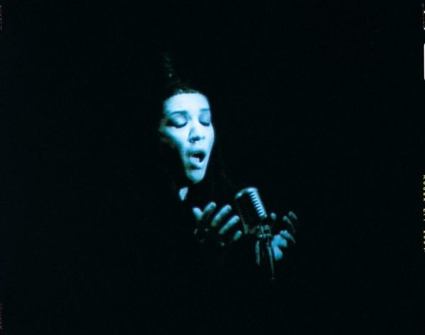 A dark vignette surrounds a person with female features cast in a blue light and passionately singing into a microphone