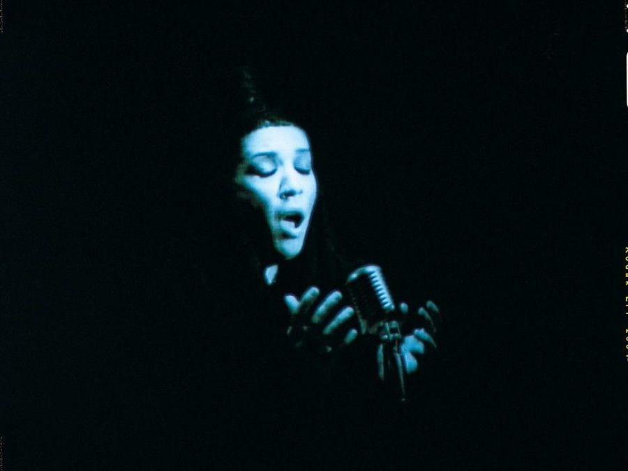 A dark vignette surrounds a person with female features cast in a blue light and passionately singing into a microphone