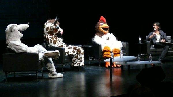 Three people dressed in animal costumes—a rat, a cow, and a chicken—sit on a stage with a fourth person.