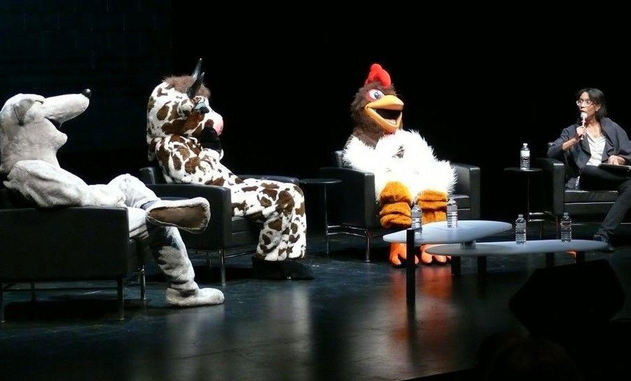 Three people dressed in animal costumes—a rat, a cow, and a chicken—sit on a stage with a fourth person.