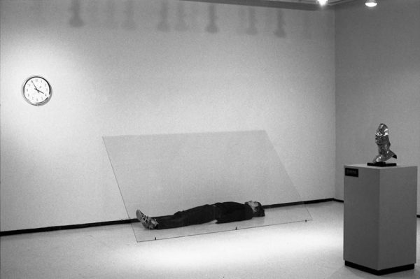 A man lying on the floor underneath a sheet of glass in a gallery with a clock on the wall to the left and a sculpture of a person's face on a pedestal to the right of the glass