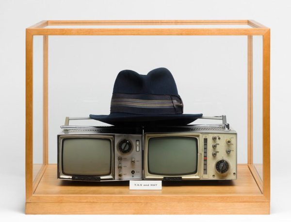 Wooden display case with a fedora atop a double cctv screen and a small white placard reading TVs and Hat