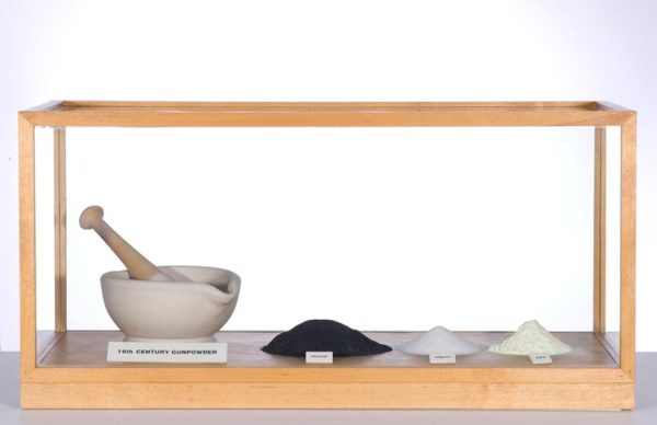 A wooden display case featuring, from left to right, a mortar and pestle, a pile of black powder, and two small piles of white powders, with labels in front of each