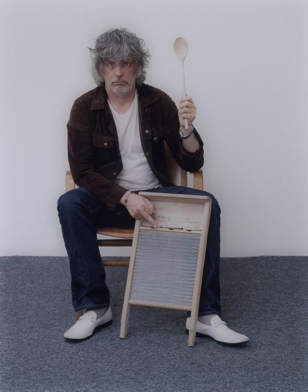 A light-skinned man with gray and disheveled chin-length hair sits on a chair, staring at the camera. In his left hand, he holds a long wooden spoon straight up in the air. His right middle finger is extended above a wood-and-metal washing board against his leg.