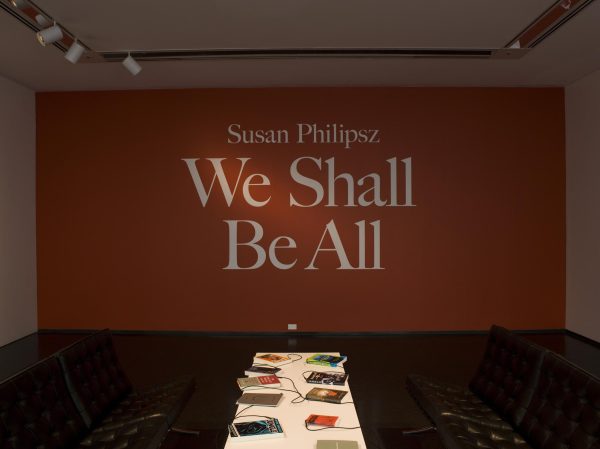 A white table with books on it and two black chairs appear in front of an orange wall with white text that reads 