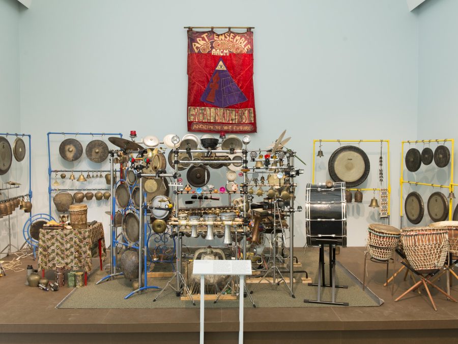 An eclectic array of percussion instruments fills a stage. Above them hangs a red banner featuring a pyramid and the words