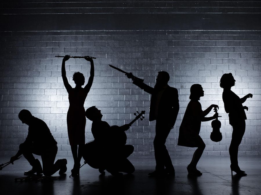 Black silhouettes of three men and three women hold instruments in unusual ways, in front of a grey brick wall.