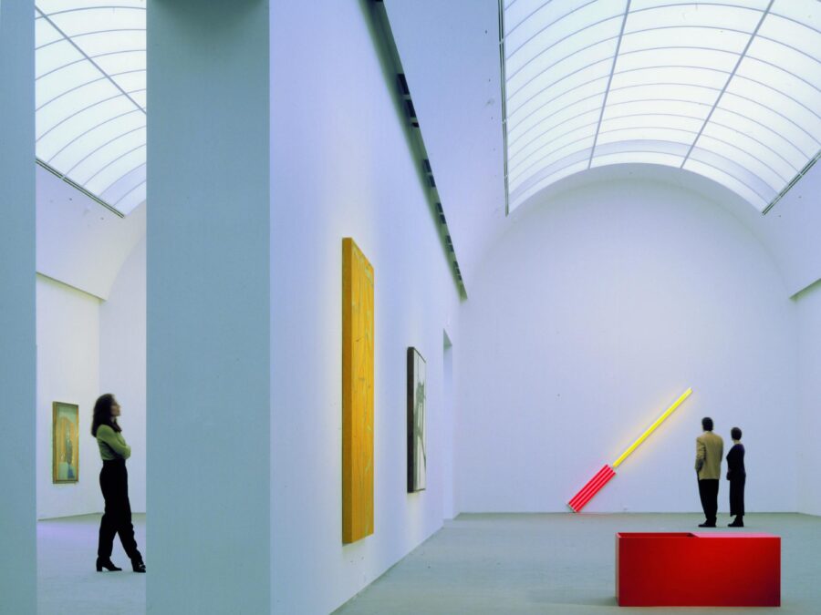 A light-filled gallery with white walls has a curved barrel vault ceiling and three visitors viewing artworks.