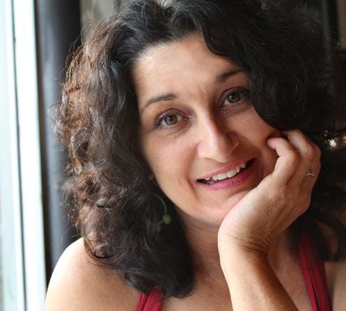 An olive-skinned woman with wavy, shoulder-length brown hair, rests her chin on her hand.