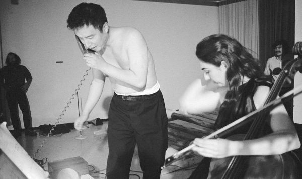A shirtless man holds a corded telephone receiver up to his hear next to a woman playing an upright bass in a room with two other people