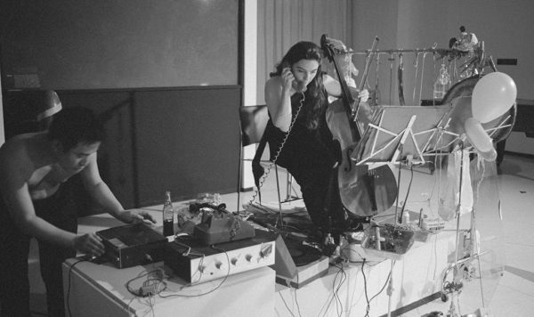 A woman holds a telephone receiver in one end and a small upright bass in the other by a table filled with sound systems and music making instruments. At the end of the table, a shirtless man works on a tape recorder.
