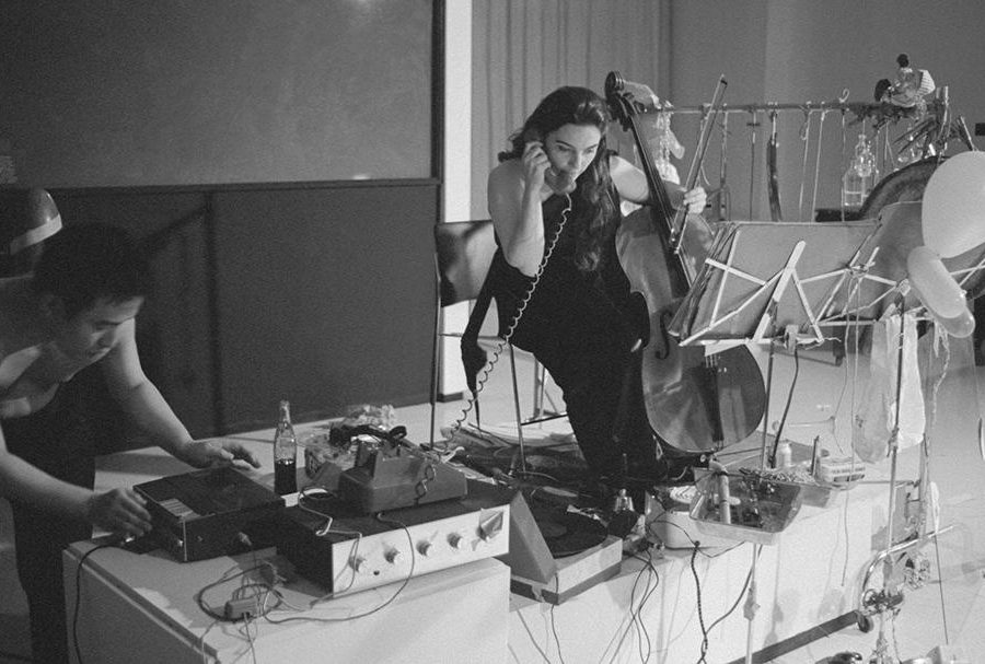 A woman holds a telephone receiver in one end and a small upright bass in the other by a table filled with sound systems and music making instruments. At the end of the table, a shirtless man works on a tape recorder.
