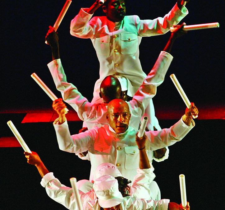 A performance view of black men and women dressed in all white sailor suits hold white cylindrical batons in both hands, sitting on top of each others shoulders while onstage. The red and green stage lighting is casted onto them.