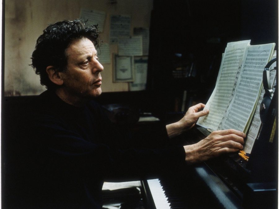 A middle-aged white man with dark brown hair dressed in all black sits at a piano in pensive thought holding pages of sheet music.