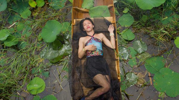 A person lays in a small wooden boat floating among lily pads and reeds