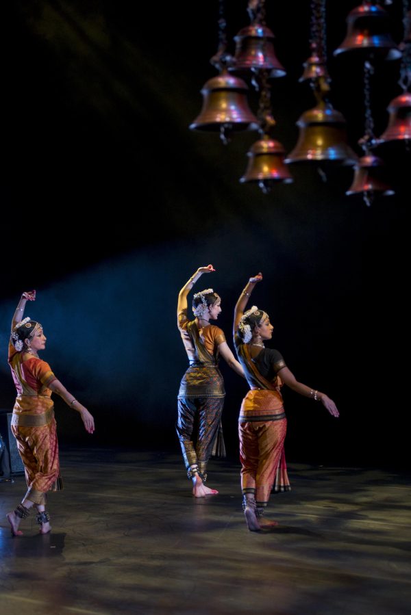 Three women wearing Indian saris, jewelry, and head ornaments dance in unison on a bare stage beneath hanging brass bells.