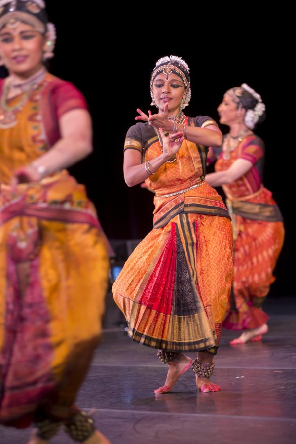 Three women on stage wearing Indian saris, elaborate head pieces, and jewelry, dance barefoot on a stage with arms bent and fingers flexed.