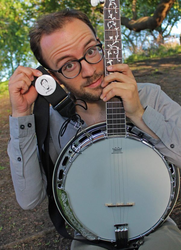A young man with round glasses and facial hair holds the neck of a bango near his face with one hand while holding a white badge with an image of a face and the word 