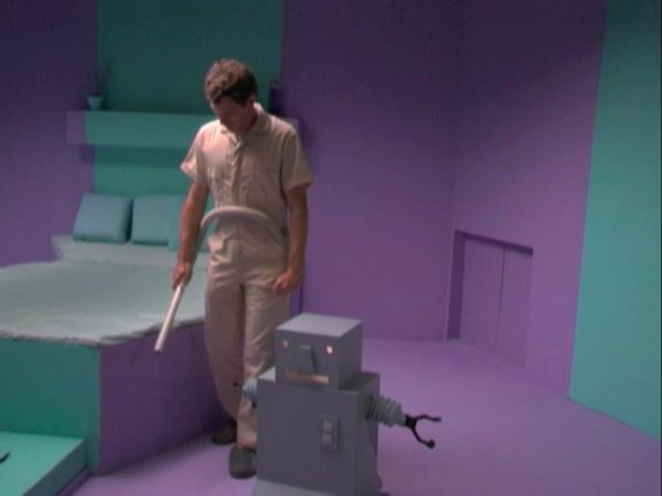 Film still of a man in a khaki jumpsuit and a grey, cubic robot in a purple and teal futuristic bedroom